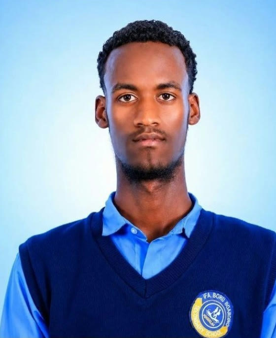 Sedem Abdulaziz photographed against a pale blue studio backdrop in his school uniform, a navy sleeveless pullover over a light blue shirt, the Ifa Boru Boarding School crest on the chest.
