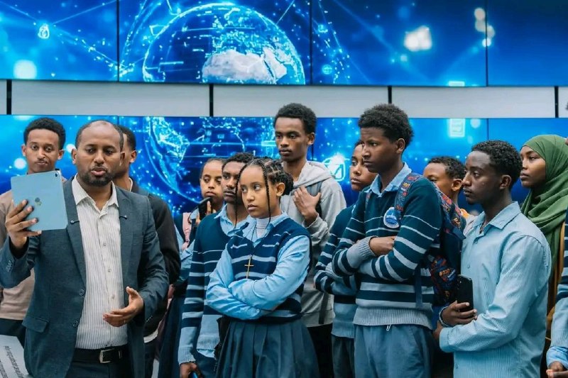 Secondary school students in uniform standing in front of large blue screens showing a globe and network graphics, while a man in a suit holds up a tablet and speaks to them during an exhibition tour.