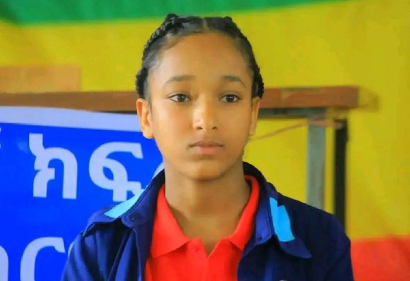 Hidase Zemenu, in a red polo shirt under a blue and light blue school tracksuit jacket, her hair braided back, seated in front of an Ethiopian flag hung on the wall behind her.