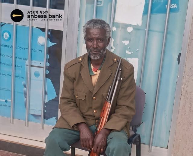 An older man with grey hair and a white beard sits on a chair outside a bank branch in a khaki uniform jacket, a rifle resting upright against his shoulder, with the Anbesa Bank sign on the window behind him.