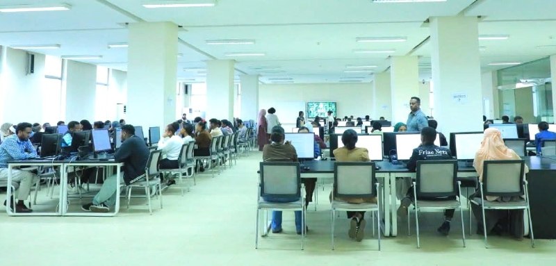 A large hall of candidates seated at desktop computers in rows, with an invigilator standing in the aisle.